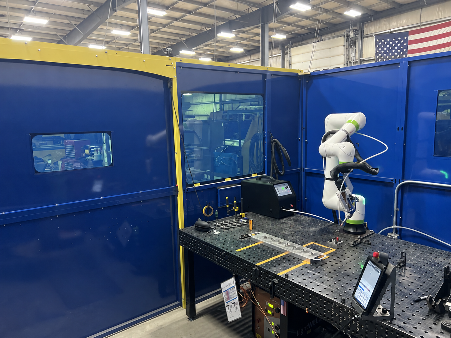 Robotic welding workstation inside a blue-and-yellow LightSAFE enclosure with laser windows and a fixture table.